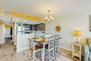 A dining area with a wooden table and chairs, a kitchen in the background, and a small side table with a lamp. - Sand Dunes Resort 2443/44 (Myrtle Beach)