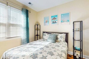 A bedroom with a bed, a window with blinds, a shelf with decorative items, and wall art. - Sun and Sand Retreat (Myrtle Beach)