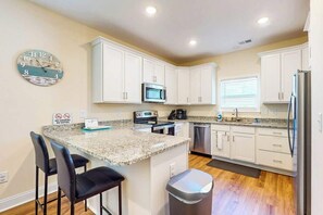 A kitchen with white cabinets, a granite countertop, a stainless steel refrigerator, and a trash can. - Sun and Sand Retreat (Myrtle Beach)