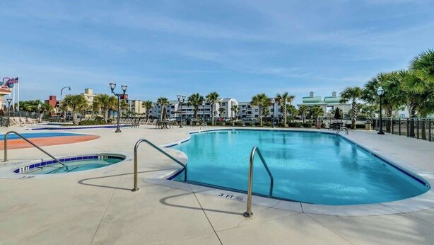 Un área de piscina con varias piscinas, incluyendo una grande en el centro y piscinas más pequeñas, rodeada por un solárium pavimentado.