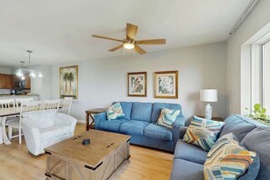 A living room with a blue sofa, a wooden coffee table, and a ceiling fan. - Tilghman Condos #1018 (North Myrtle Beach)