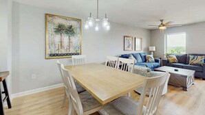 Un comedor con mesa de madera y sillas blancas, un sofá azul con cojines estampados, un ventilador de techo y un cuadro grande en la pared.