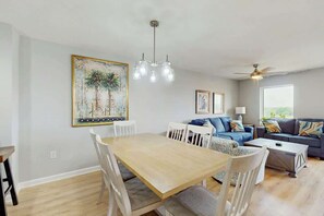 A dining area with a wooden table and white chairs, a blue sofa with patterned cushions, a ceiling fan, and a large framed painting on the wall. - Tilghman Condos #1018 (North Myrtle Beach)