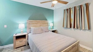 A bedroom with a wooden bed, two bedside tables with lamps, a ceiling fan, and a wall with curtains. - Tilghman Condos #1217 (North Myrtle Beach)