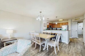 A dining area with a wooden table and chairs, a kitchen in the background, and a door leading to another room. - Tilghman Condos #1217 (North Myrtle Beach)