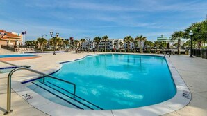 A swimming pool with a clear blue water, surrounded by a paved deck, palm trees, and a building with a green roof in the background. - Tilghman Condos #1217 (North Myrtle Beach)