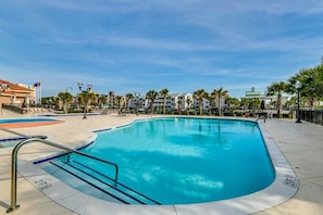 Ein Swimmingpool mit klarem, blauem Wasser, umgeben von einer gepflasterten Terrasse, Palmen und einem Gebäude mit grüner Dachbegrünung im Hintergrund.
