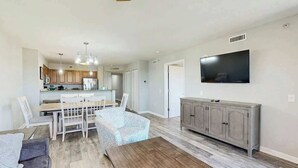 A spacious living area with a dining table and chairs, a sofa, a TV mounted on the wall, and a wooden bench. - Tilghman Condos #1217 (North Myrtle Beach)