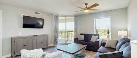 A living room with a flat-screen TV, a ceiling fan, a sofa, a coffee table, and a window with a view.