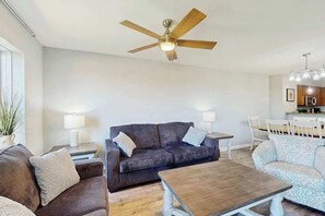 A living room with a dark brown sofa, a wooden coffee table, and a ceiling fan. - Tilghman Condos #1217 (North Myrtle Beach)