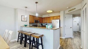 A modern kitchen with wooden cabinets, a central island with stools, a microwave, and a refrigerator. - Tilghman Condos #1217 (North Myrtle Beach)