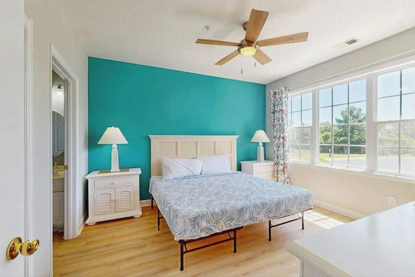 A bedroom with a bed, two bedside tables, a ceiling fan, and a window with a view. - Palmy Days at Ironwood 424 (North Myrtle Beach)