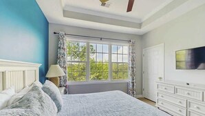 Ein Schlafzimmer mit einem großen Bett, einem Fenster mit Vorhängen, einem an der Wand befestigten Fernseher und einer weißen Kommode.