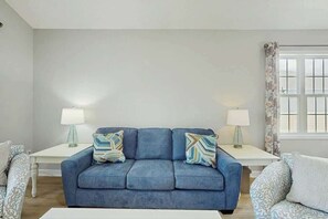 Un salón con un sofá azul, dos mesitas de noche con lámparas y un ventanal con cortinas.