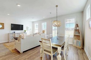 A modern living room with a sofa, dining table, and chairs. There is a television on the wall and a decorative light fixture above the table. - Double Eagle Unit 15 (North Myrtle Beach)