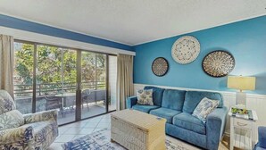 A living room with a blue sofa, a wicker coffee table, and a patterned rug. There are decorative wall plates and a lamp on a side table. - Ocean Creek Unit 2359 (Myrtle Beach)