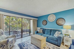 Ein Wohnzimmer mit einem blauen Sofa, einem Korbtisch und einem gemusterten Teppich. An den Wänden hängen Wandteller und auf einem Beistelltisch steht eine Lampe.