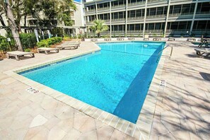 Piscina retangular com borda azulejada, cercada por espreguiçadeiras e um deck pavimentado.