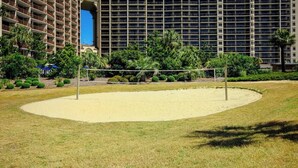 A sports court with a volleyball net, surrounded by a grassy area and a building with multiple balconies in the background. - Ocean Creek Unit 2359 (Myrtle Beach)