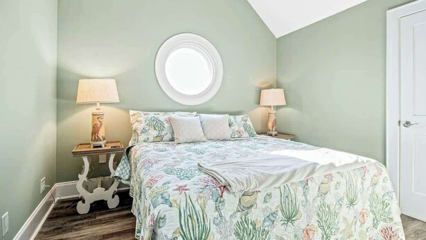 A bedroom with a bed, two bedside tables with lamps, a round window, and a door. - Hermosa Villa Retreat (North Myrtle Beach)