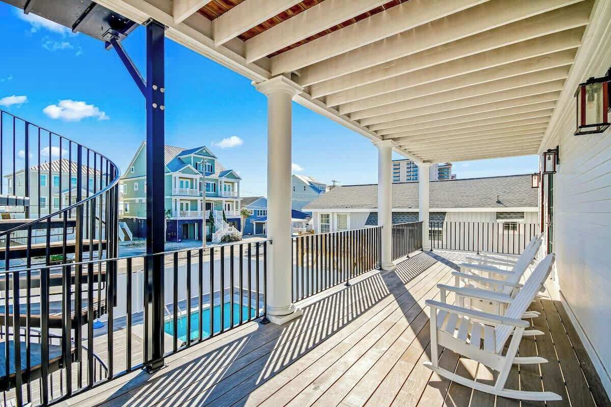 Um deck coberto ao ar livre com piscina, cadeiras de balanço brancas e vista para casas ao fundo.