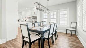 A modern kitchen with white cabinets, a dining table with chairs, and a hanging light fixture. - Hermosa Villa Retreat (North Myrtle Beach)