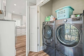 Une buanderie équipée de deux lave-linge et deux sèche-linge à chargement frontal en acier inoxydable, un panier à linge bleu posé sur le dessus, et une porte blanche donnant sur un couloir.