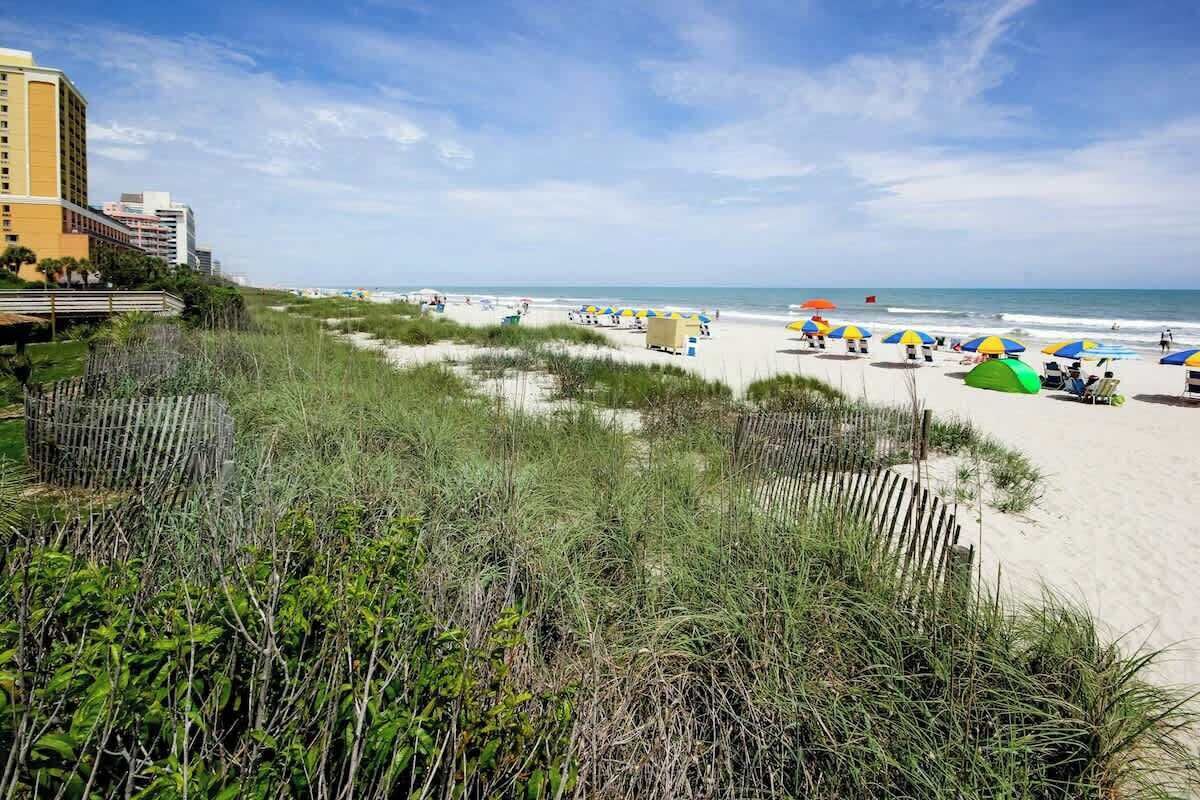 A beach with a hotel in the background, beach umbrellas, and sand dunes.