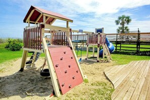 A wooden play structure with a slide and climbing area, set on sandy ground with a fence in the background. - Beach & Golf Vacation Dream (Myrtle Beach)
