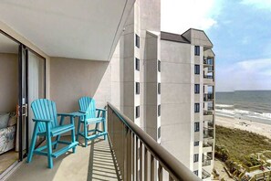 A balcony with two chairs and a table, overlooking a beach and a high-rise building. - Sea Winds Unit 608 Condo (North Myrtle Beach)
