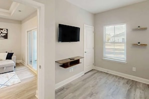 Ein Zimmer mit flachbildfernseher an der Wand, einer Schiebetür, einem Fenster mit Jalousien und einem kleinen Holzregal.