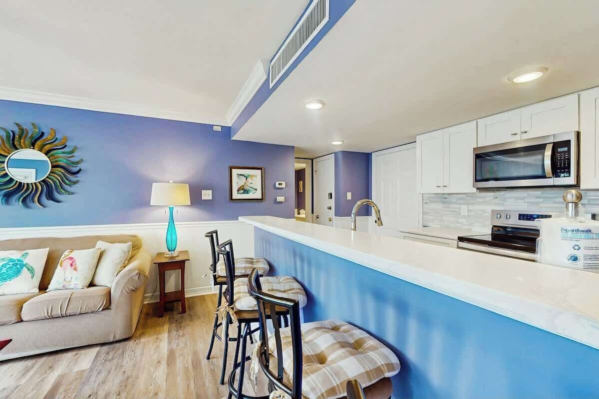 A modern kitchen with white cabinets, a microwave, and a built-in oven. There is a bar area with stools and a small table with a lamp.