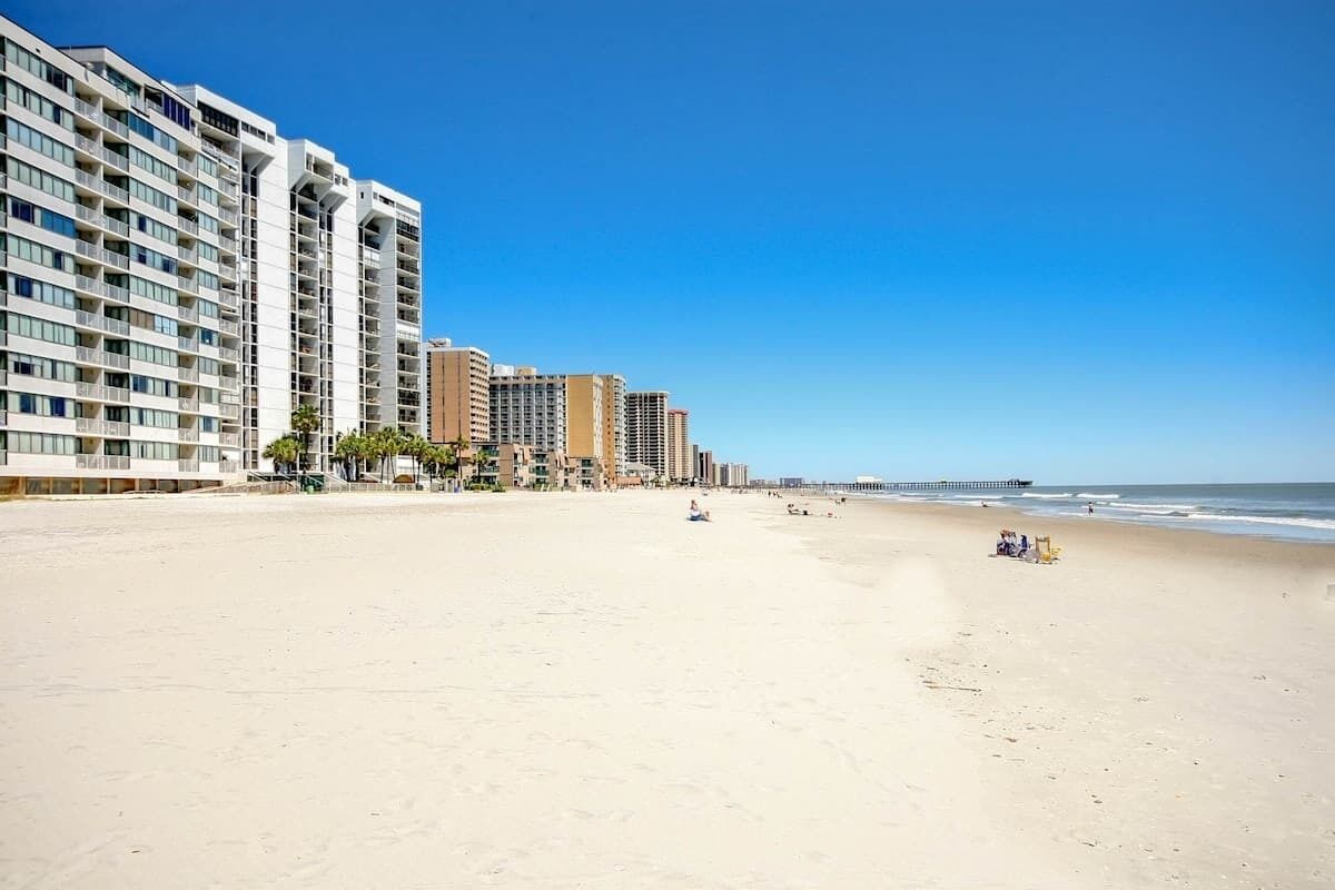 A beachfront hotel with multiple floors, a sandy beach, and a clear blue sky.