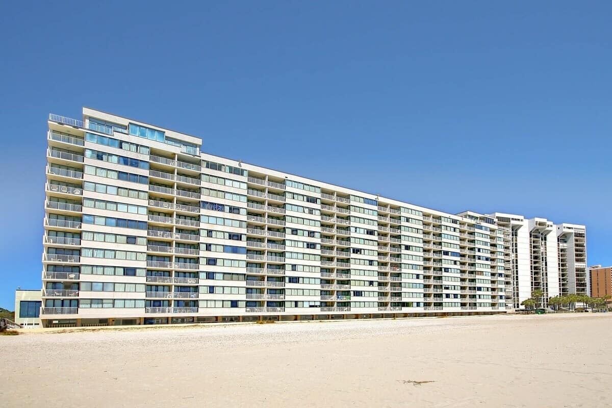 A multi-story hotel with balconies, located near a beach.