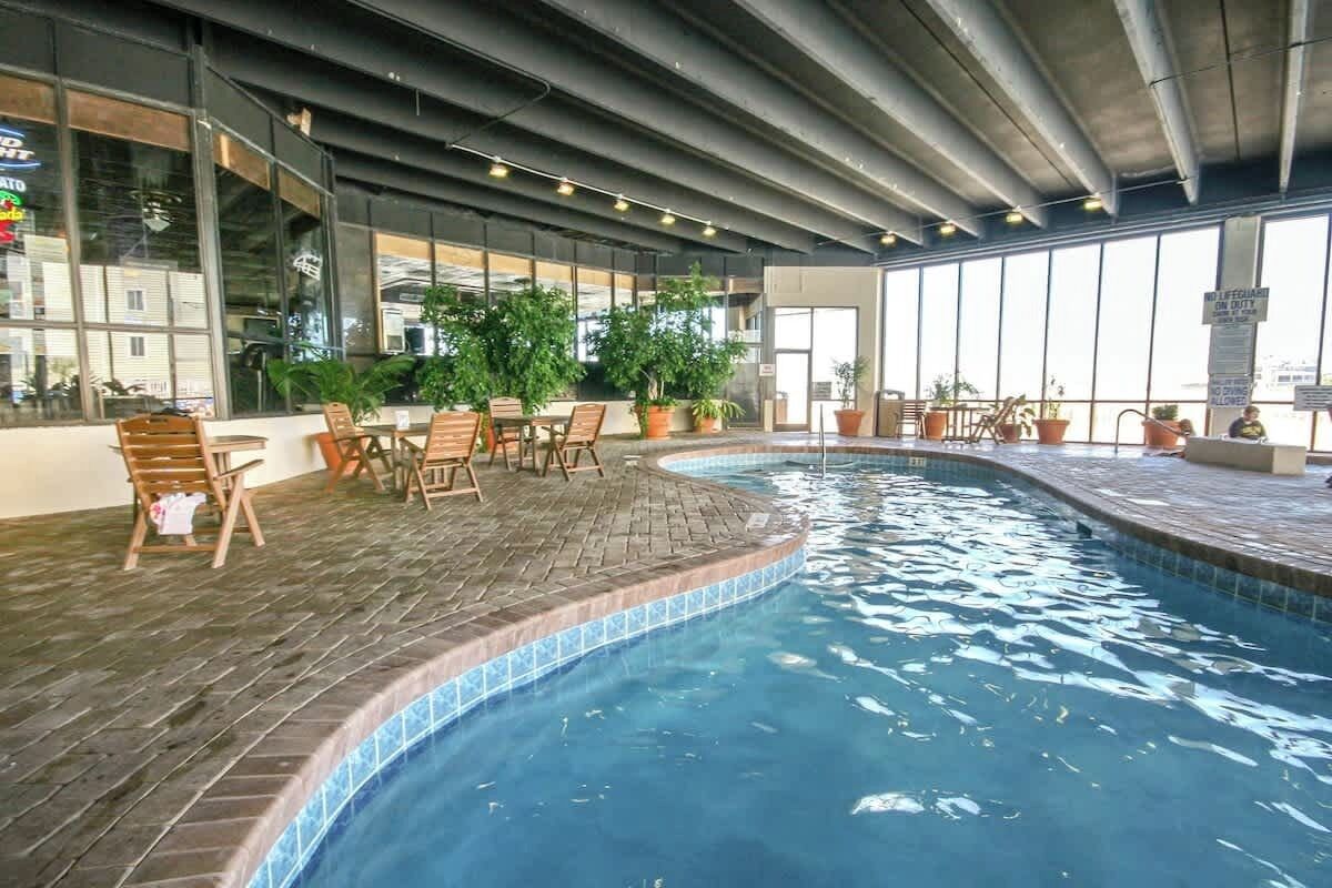 An indoor swimming pool with a tiled edge, surrounded by wooden chairs and potted plants.
