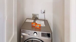 A compact, stainless steel front-loading washing machine with a control panel, a power outlet, and a small shelf with Tide detergent boxes. - River Oaks Unit 61M Condo (Myrtle Beach)