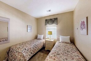 Ein Schlafzimmer mit zwei Einzelbetten, einem Nachttisch mit Lampe, einem Fenster mit Jalousien und einem an der Wand befestigten Spiegel.