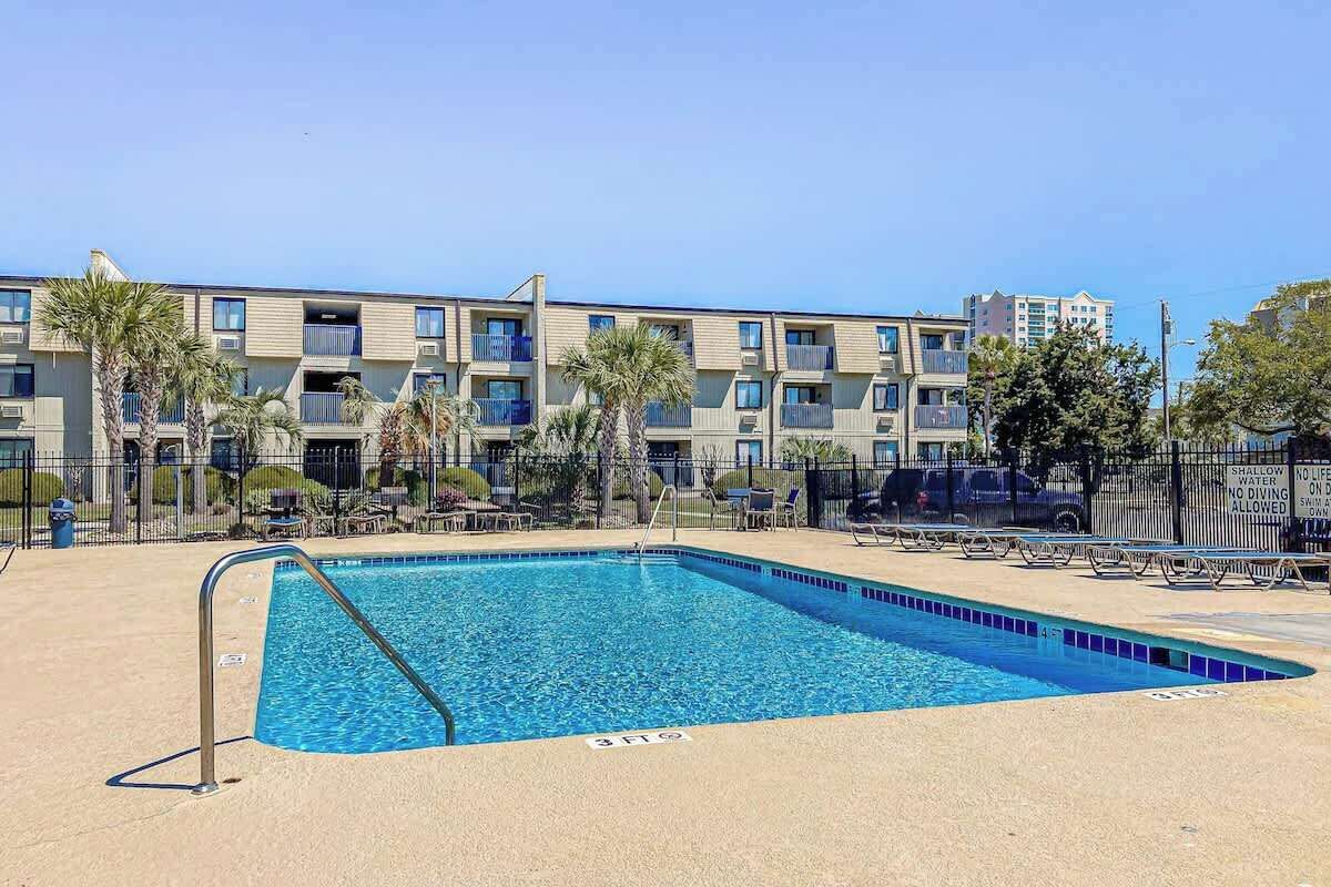 Piscina com escada de metal, cercada por uma grade e palmeiras, com um edifício de vários andares ao fundo.