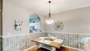 Uma área de refeições com mesa e cadeiras de madeira, luminária pendente e um compasso decorativo fixado na parede.