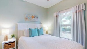 Quarto com cama, luminárias de cabeceira, mesa de cabeceira, janela com cortinas e uma obra de arte na parede.