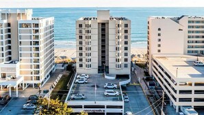 Un hotel de varios pisos con estacionamiento frente a él, ubicado cerca de una playa y el océano al fondo.