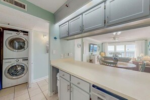 A kitchen with two stacked washing machines, a dining area with a round table, and a view of the living room. - Sea Marsh II Unit 1002 (North Myrtle Beach)