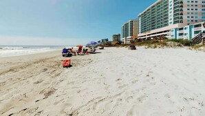 Una playa con un edificio de hotel, sillas de playa, sombrillas y cielo despejado.