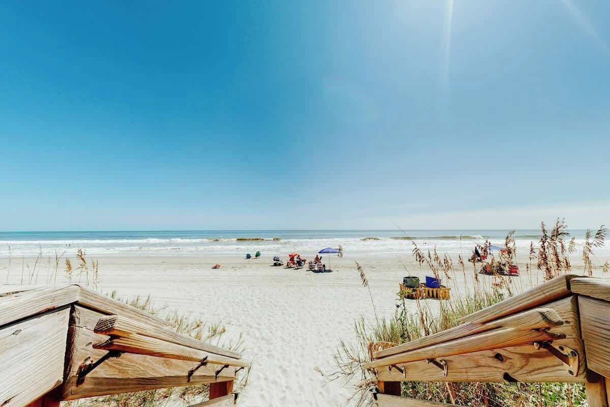 Une plage avec des structures en bois, peut-être une maison de plage ou un complexe hôtelier, avec un ciel dégagé et l’océan en arrière-plan.