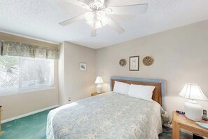 A hotel room with a bed, a ceiling fan, a window with blinds, a bedside table with a lamp, and a wall clock. - Ocean Garden Villas G-2 (North Myrtle Beach)