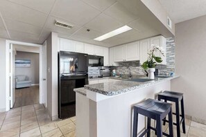 Une cuisine moderne avec des armoires blanches, un comptoir en granit, un réfrigérateur en acier inoxydable et un four encastré. On y trouve deux tabourets de bar et une plante en pot sur l’îlot central.