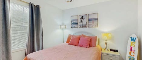 Un dormitorio con cama, mesita de noche, lámpara, cartel de tabla de surf y ventana con cortinas.
