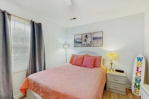 A bedroom with a bed, a nightstand, a lamp, a surfboard sign, and a window with curtains. - Hawleywood Too Retreat (Surfside Beach)