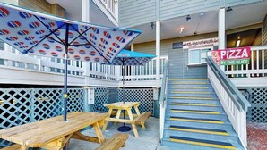 Une table et des bancs de pique-nique en bois, placés sous un parasol à l’effigie de Pepsi, une enseigne de pizzeria et un escalier orné de bandes de sécurité jaunes.