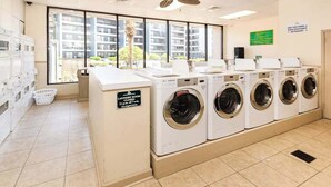 Une buanderie équipée de plusieurs machines à laver, un panneau indiquant que la pièce est fermée, et une vue sur un immeuble d’habitation à travers de grandes fenêtres.